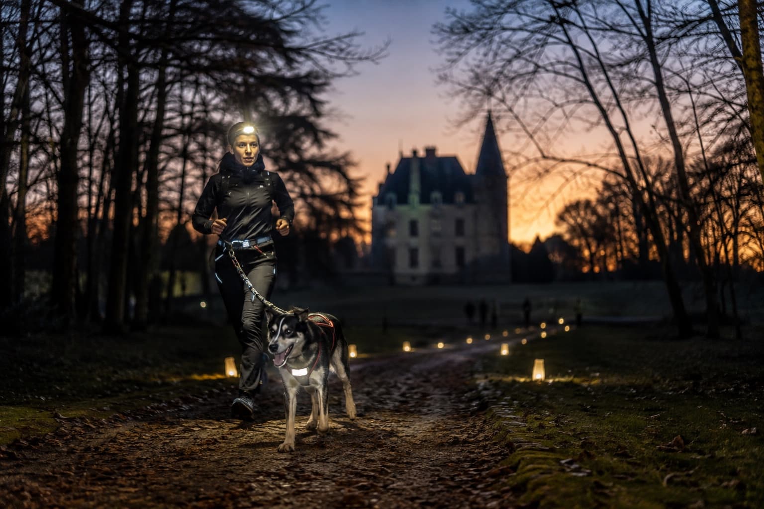 Coureuse avec lampe frontale et chien en harnais sur un sentier au crépuscule devant un château